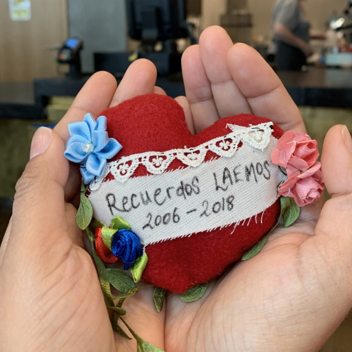 Two hands caressing a small red heart with Recuerdos LAEMOS written on a white cloth, surrounded by fabric flowers.