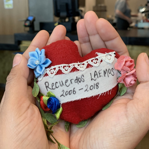 Two hands caressing a small red heart with Recuerdos LAEMOS written on a white cloth, surrounded by fabric flowers.
